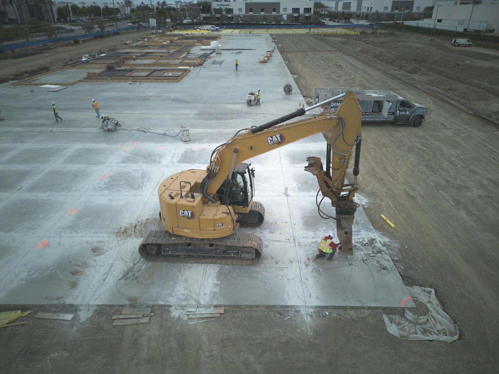 Aerial drone view of DSI CAT excavator with hoe ram breaking large commercial concrete slab with DSI truck on site